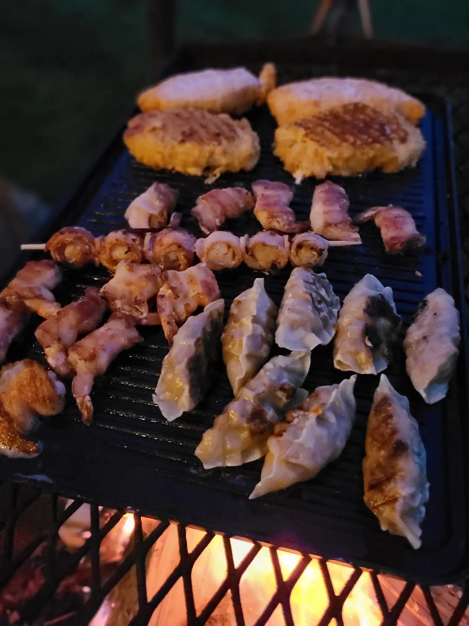Grilled food items including skewers and dumplings on a griddle on an anvil grill with flames below.