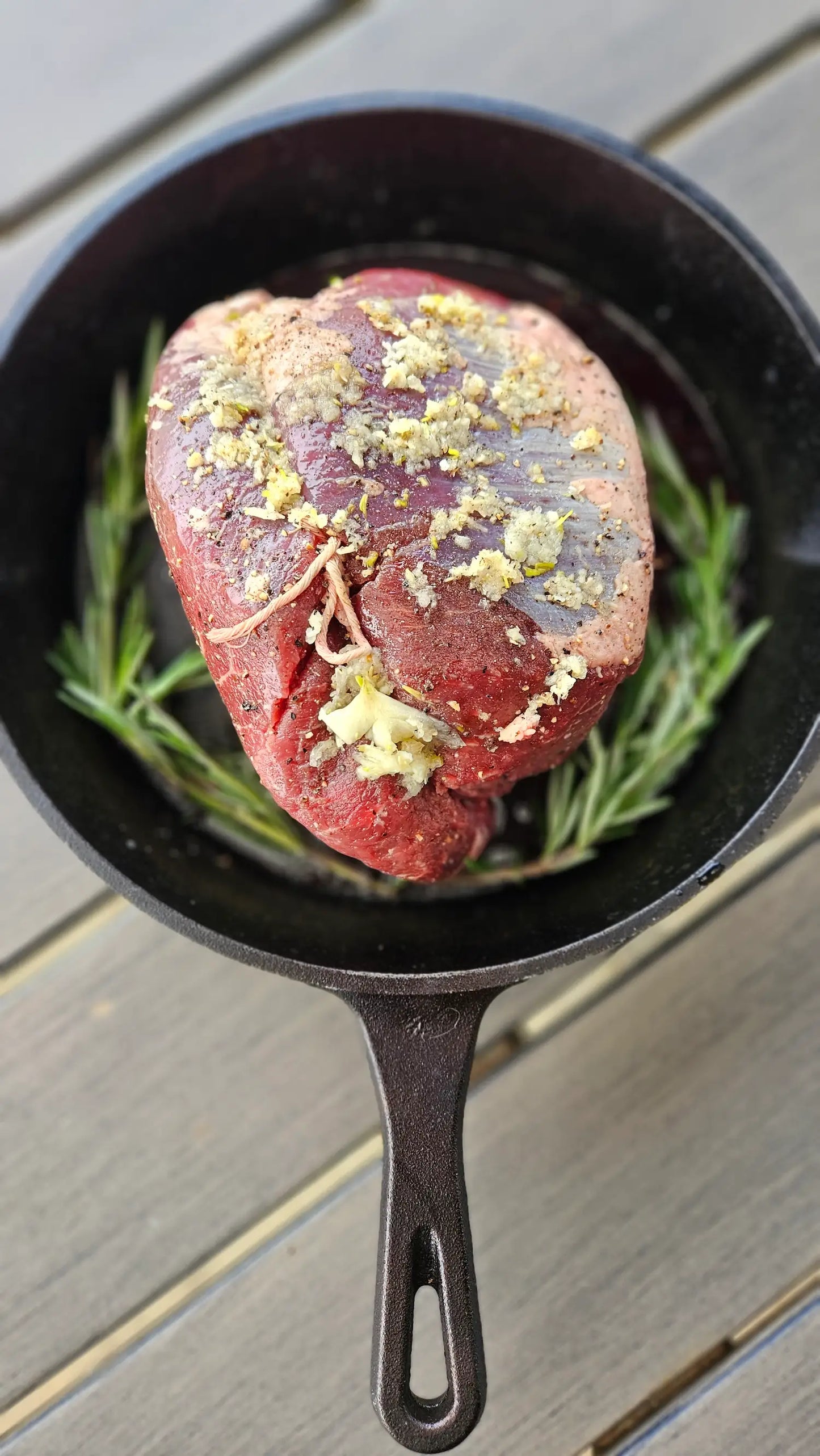 Raw beef tenderloin with garlic and rosemary in a cast iron skillet on a wooden surface