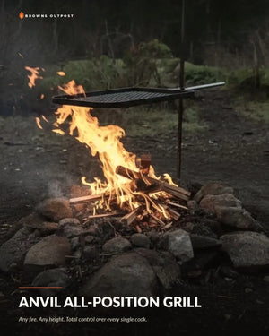 Anvil all-position grill shown over an outdoor campfire with rocks and wood, with flames and a hanging metal grate visible. Text at the top reads 'Browns Outpost' and the bottom reads 'ANVIL ALL-POSITION GRILL' with a tagline.